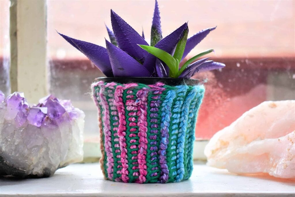 Tunisian crochet ribbing cosmic crochet coozies - a amethyst crystal an aloe plant and a rose quartz in a window sill, the plant has a crochet rainbow coozie