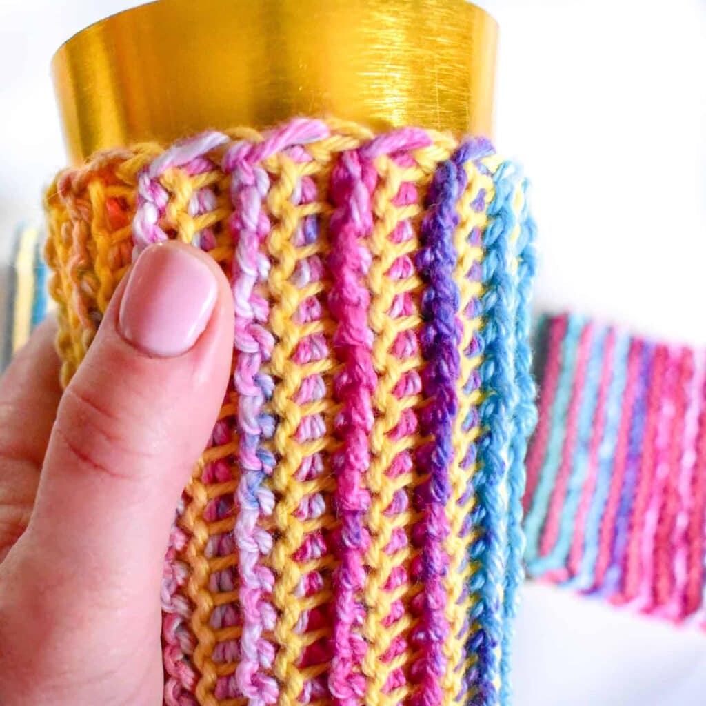 tunisian crochet ribbing pattern a womans hand holding a rainbow crochet coozie in a yellow cup with pink nailpolish