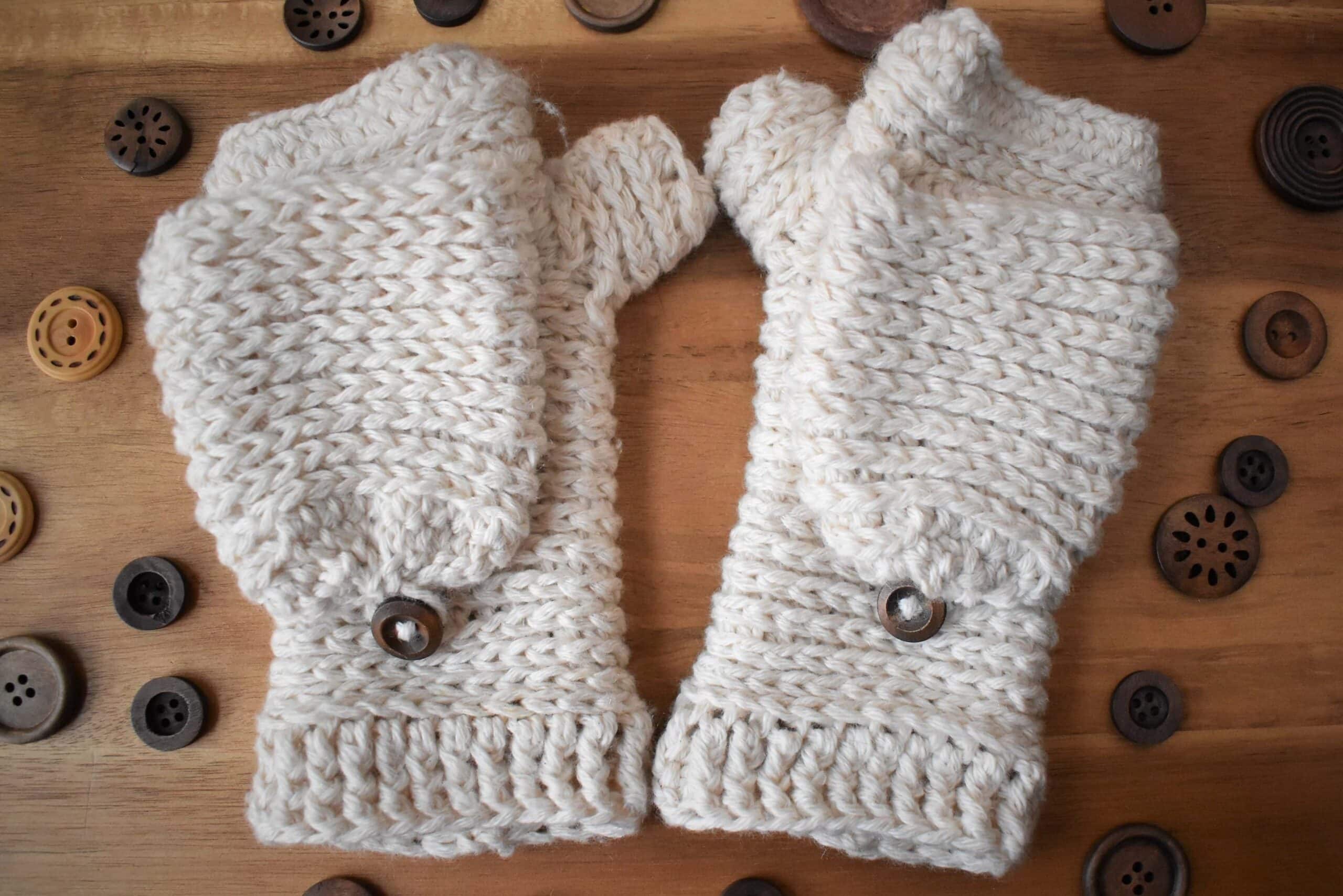 White crochet mittens on a wooden table with wood buttons scattered throughout.  Beautiful easy crochet mittens pattern