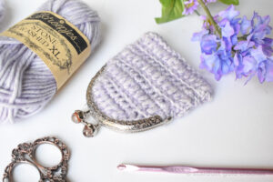 different angle of crochet coin purse lilac dreams on white table purple flowers and a crochet hook and a pair of scissors and a skein of sheepjes yarn