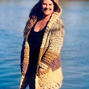 tasha margette standing in front of a lake with arms open wearing her crocheted kirra cardigan pattern