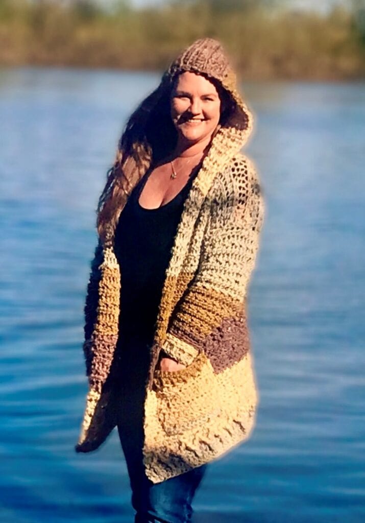 tasha margette standing in front of a lake with arms open wearing her crocheted kirra cardigan pattern